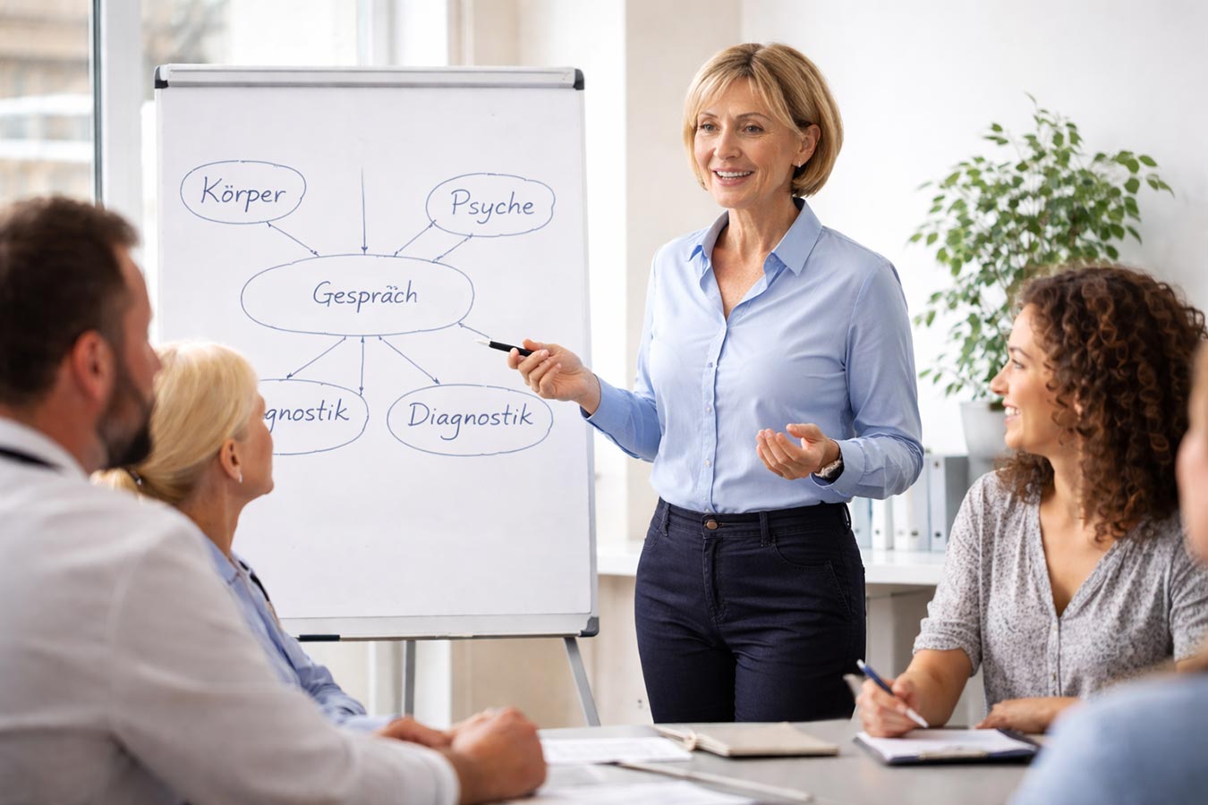 Weiterbildungsteilnehmer hören der Dozentin zu, die vorne im Raum an einer Flipchart steht - Symbolbild Ausbildung Psychosomatische Grundversorgung